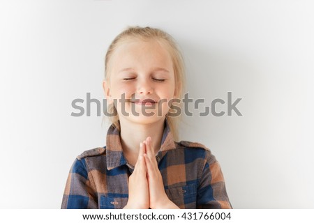 Pretty little girl with closed eyes smiling and meditating with happy face in morning light. Blond Caucasian kid putting hands together as if praying for lovely day to come or making wish for birthday
