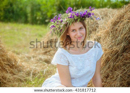 Young beautiful blonde girl in a white dress and a wreath of wild flowers sitting near haystack. Summer in the village