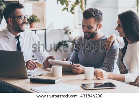 Just put your signature here! Cheerful young man signing some documents while sitting together with his wife and man in shirt and tie  Royalty-Free Stock Photo #431846200