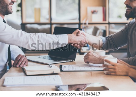 Sealing a deal. Side view close-up of two young man shaking hands while sitting at the wooden desk Royalty-Free Stock Photo #431848417