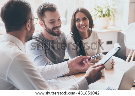 Just put your signature here! Confident young man in shirt and tie holding some document and pointing it while sitting together with cheerful young couple at the desk in office  Royalty-Free Stock Photo #431851042