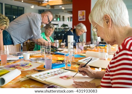 Woman in striped red and white shirt working on canvas while painting with brush at table with other students and teacher in spacious studio Royalty-Free Stock Photo #431933083