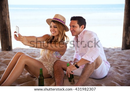 Attractive young couple taking a selfie with a smartphone while enjoying their vacation at the beach, relaxing and drinking some beer together