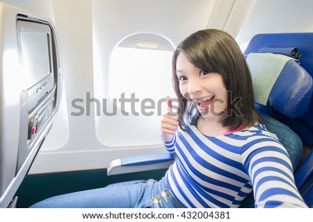 Happy young woman is sitting in the airplane and take a selfie, show thumb up, asian