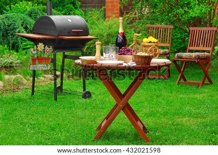 Weekend BBQ Family Party Scene On The Backyard In Summertime. Charcoal Barbecue Grill Appliance With Welcome Sign And Outdoor Wooden Furniture  On The Garden Lawn. lunch Table With Food And  Beverage