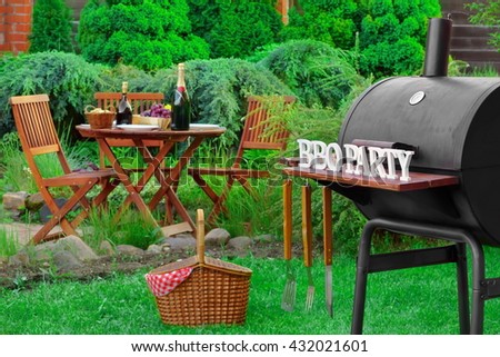 Barbecue Charcoal Grill Appliance With Tools And BBQ Party Sign, Picnic Basket And Wooden Table With Food And  Beverages On The Backyard Lawn In The  Background