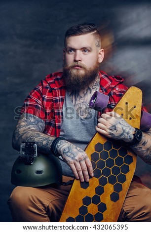 Tattooed bearded male in red shirt posing with longboard in studio.