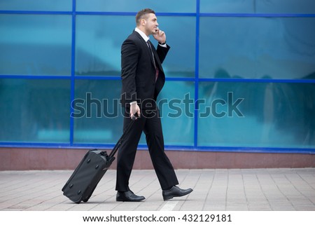 Profile view portrait of cheerful young man on business trip walking with his luggage while talking on smartphone in front of modern glass building outdoors. Travelling guy making call. Copy space Royalty-Free Stock Photo #432129181