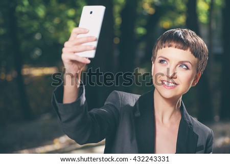Outdoors portrait of Attractive young short hair smiling woman in forma lwear taking photo with her cellphone in park