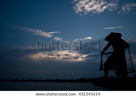 Silhouette of asian fisherman on wooden boat in action casting a net for catching freshwater fish in nature river in the early morning before sunrise