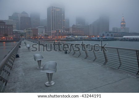 San Francisco cityscape with Ferry Terminal at down shot from Pier 14 with metal swivel chairs, top of buildings disappearing in fog and low clouds before winter sunrise