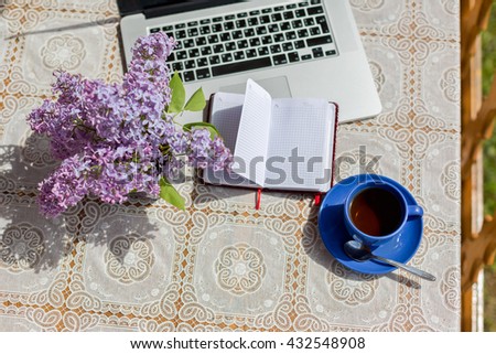 Open Notepad and blue cup of tea next to the laptop and vase with lilacs on the table with a white tablecloth with space for text