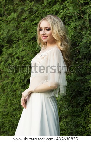 Beautiful girl in fantasy ball gown with thuja green fence in the background. Pretty young woman in wedding dress with evening makeup and natural hairstyle. 