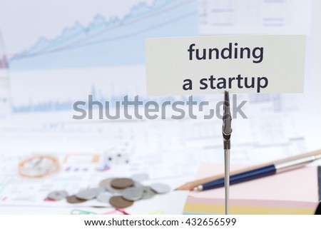funding a startup- a message on the photo holder with background of  stationary, money, compass,  and stock chart 
