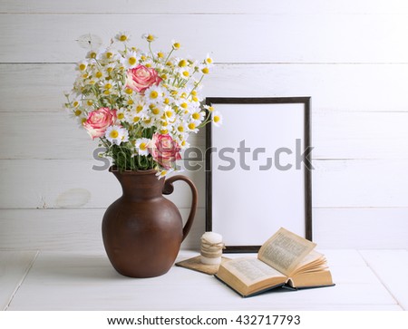 Daisy bouquet with roses in clay jug with open book and motivational frame  for your text or picture on background of white wooden planks in scandinavian style. Home interior. 
