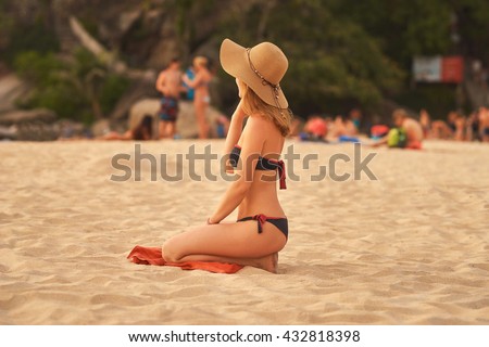 Young woman with a big hat sits on the sandy beach during the sunset  and looks into the distance 