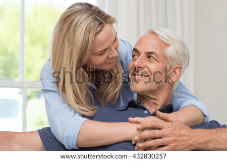 Portrait of a loving mature senior woman embracing man from behind at home. Happy senior couple relaxing at home while wife hugging husband. Smiling woman hugging her retired husband at home.
