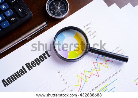 Magnifying glass on colourful pie chart with "Annual Report" text on paper, dice, spectacles, pen, laptop calculator on wooden table - business, banking, finance and investment concept