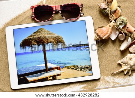 high-angle shot of a white wooden surface full of sand and an ornament made with different conchs and seashells, a pair of sunglasses and a tablet computer with the picture of a beach taken by myself