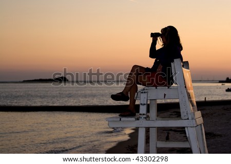 A silhouette of a woman looking with binoculars at the beach while sitting on a lifeguard chair.