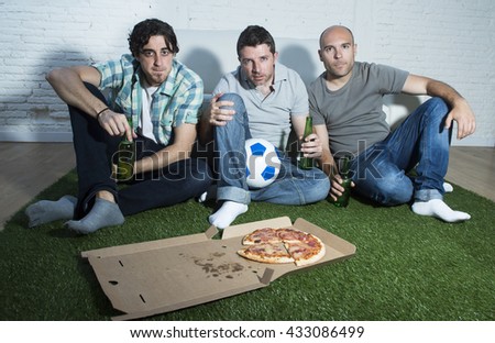 group of friends fanatic football fans watching soccer game on television with beers and pizza on grass carpet emulating stadium pitch looking nervous and stressed very focused on the match