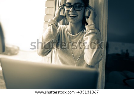 Young female at home.She sitting in her living room by the window and listening to music on laptop.