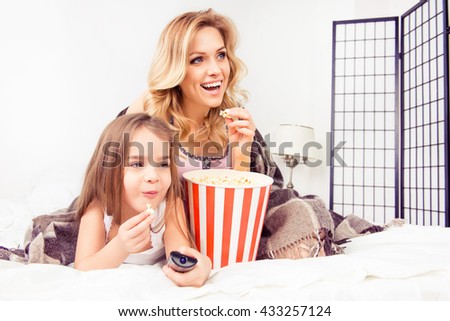 Cheerful smiling little girl and her mom watching tv with popcorn