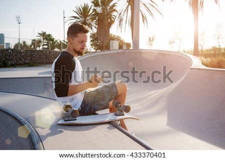Young man is holding a mobile phone while sitting in a skate zone. A guy is surfing the web by the smart-phone while sitting on a sunset background. A photo with flare light and sunny background.