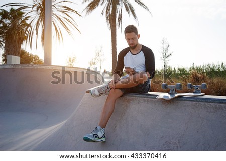 Young guy is checking the email while having break in a skate park on a sunny summer day. Skateboarder is holding a mobile phone while sitting in a skate zone. Flare light.
