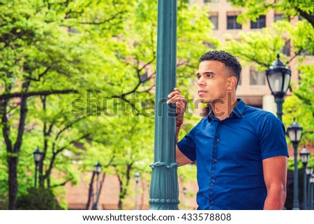 African American college student, wearing blue short sleeve shirt, standing by light pole on street on campus in New York in summer, looking away, thinking, lost in thought. Instagram filtered effect.