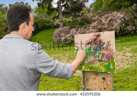 Side view of a male artist painting with oils and acrylics finishing touches working  on a trestle and easel  during an art class in a park