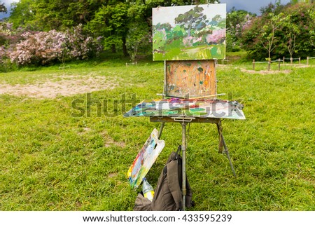 Painter's sketchbook with multicolored  palette of blended oil paints and professional paintbrushes on the tripod in a garden