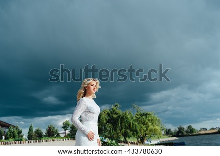 Beautiful young bride woman in luxury wedding dress with red lips makeup and long wavy hairstyle. Beauty girl portrait posing against the background of dark rainy clouds, outdoor photo.