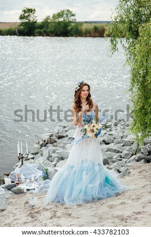 Beautiful young bride woman in luxury wedding dress with red lips makeup and long wavy hairstyle. Beauty girl portrait posing near water, outdoor photo.