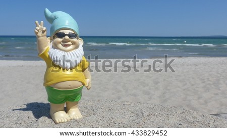 Garden gnome on vacation at sea  makes  peace sign. He stands on the sandy beach in front of the blue ocean with waves and cloudless sky. Baltic Sea, Schleswig-Holstein, Germany, Europe