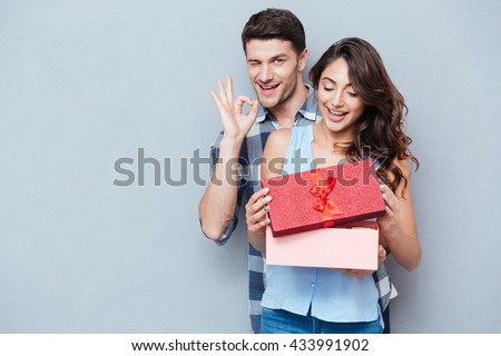 Young beautiful woman receiving a gift from her boyfriend showing ok sign isolated on gray background