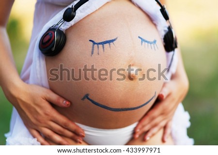 Prenatal education. Pregnant woman belly closeup with smiling funny face drawing listening to the music outdoors. Earphones at woman's belly. Happy pregnancy, fun, creativity.
