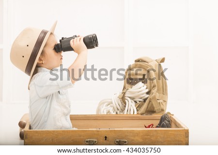 Adorable little explorer girl in a safari hat and explorer clothes with binoculars playing Safari sitting in a old wooden suitcase. Little child playing treasure hunt. Looking for the summer vacation Royalty-Free Stock Photo #434035750