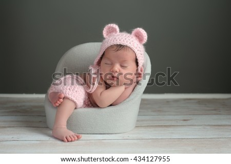 A two week old newborn baby girl sleeping in a little chair. She is wearing a crocheted, pink bear bonnet and matching shorts. Shot in the studio on a gray background. Royalty-Free Stock Photo #434127955