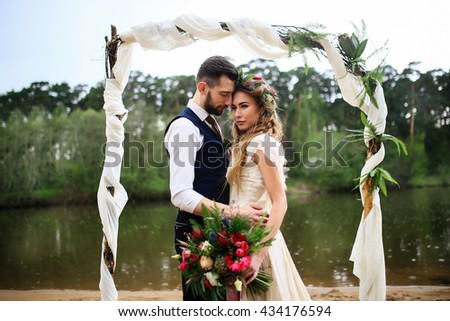 the groom in a wedding suit and boutonniere and bride in lace dress with bouquet and wreath , wedding arch on a background of lake Royalty-Free Stock Photo #434176594