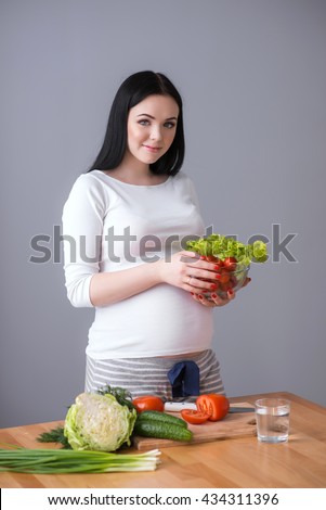 Beautiful pregnant woman looking at camera and smiling standing at the table. The happiest time for every woman. Vegetables and food in the hands of.