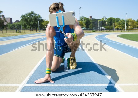 Athlete crouching at the starting line of a running track taking selfie with his mobile phone on a selfie stick