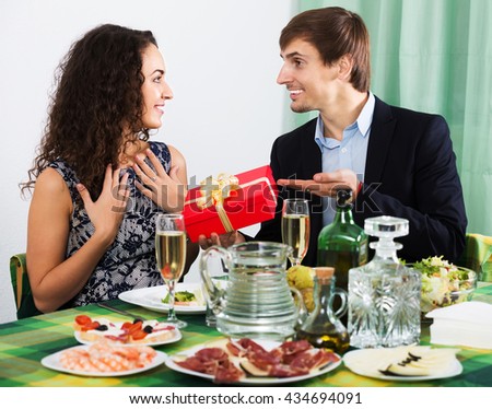Man presenting gift to positive woman during romantic dinner with champagne in home. Focus on man