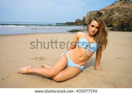 Beautiful young brunette in blue bikini lying on the sand at the beach.  She is petite, has green eyes, and has a tattoo on her left forearm.