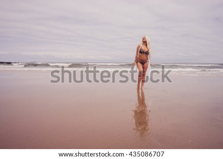 Beautiful young blonde haired model stood on the beach, the sea behind her.  She has blue eyes, wears a leopard print bikini, a cardigan.  Her reflection is captured in the sand.