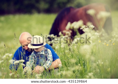Cute little boy in straw hat and his bald handsome father are examining different herbages growing on the field with a grazing horse on a background. Image with selective focus and toning