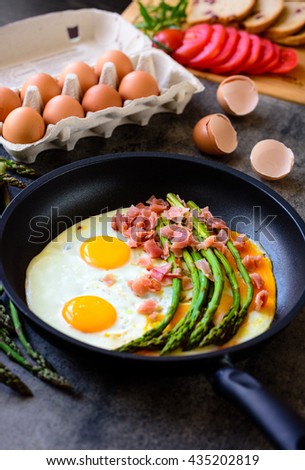 Fried eggs with asparagus, ham, eggs in container, egg shell and tomatoes on a dark stone board