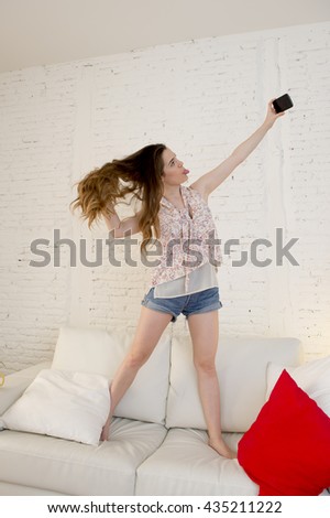 young attractive girl playing standing on top of on home sofa couch taking selfie portrait with mobile phone having fun laughing smiling happy and playful and making funny faces