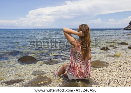 The woman and the sea. Young red hair woman sitting by the sea on the beach. Staring at sea picture. Meditation by sea image. Ecotourism illustration: clear water and sand empty beach. Loose hair girl