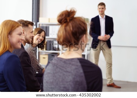 Smiling brown haired woman listening to coworkers during meeting with presenter standing in background with white bulletin board behind him Royalty-Free Stock Photo #435302002